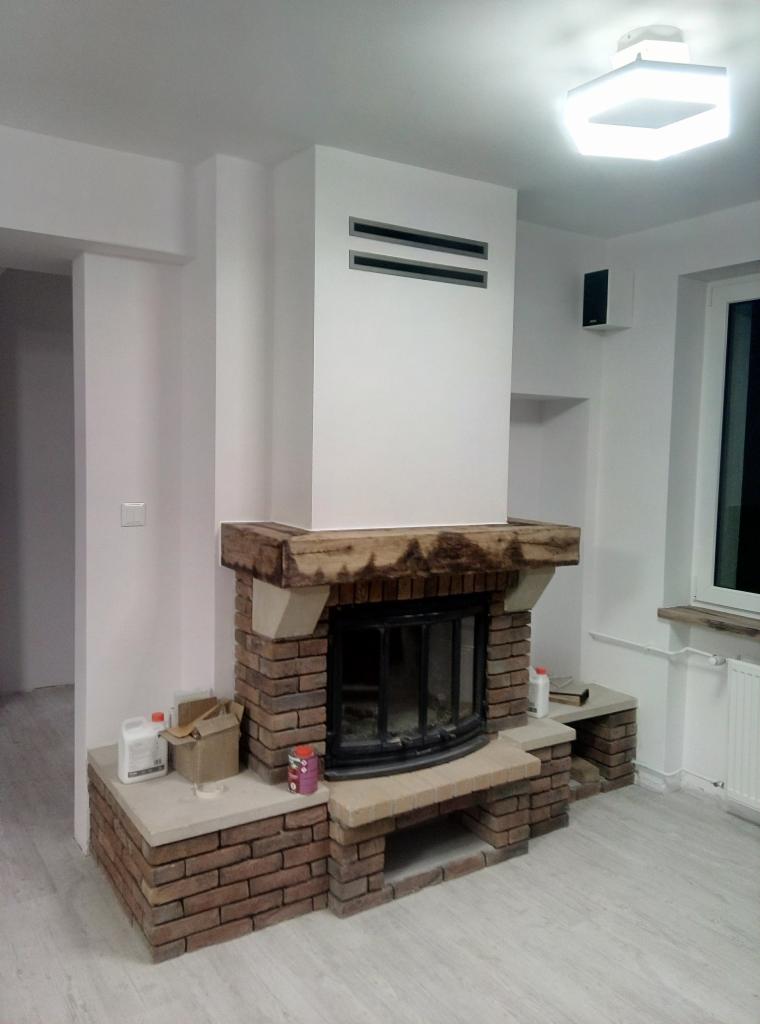 Murowany kominek z drewnianą belką nad paleniskiem, z widocznymi materiałami budowlanymi obok, w jasnym pomieszczeniu z białymi ścianami i nowoczesnym oświetleniem sufitowym.