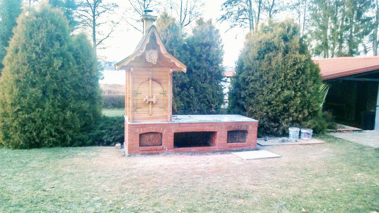 Murowany grill ogrodowy z drewnianą nadbudówką w stylu rustykalnym, otoczony tujami i trawnikiem. Widoczne dwa wiadra z farbą obok.