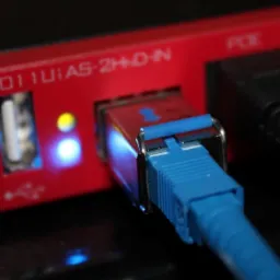 Czerwony router RB2011UiAS-2HnD-IN z podłączonym niebieskim kablem Ethernet, diody LED świecą na żółto i niebiesko, obok wpięte inne przewody.