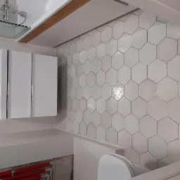 Biała szafka łazienkowa z lustrem nad umywalką, ściana wyłożona heksagonalnymi płytkami, widoczne rury instalacji wodnej.