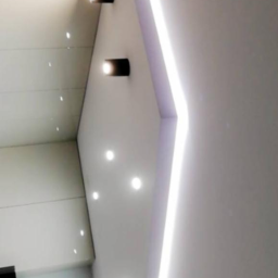 Rb-Invest - Biały sufit z oświetleniem LED w postaci listwy i punktowych reflektorów, widok z dołu, częściowo widoczny fragment zabudowy meblowej.