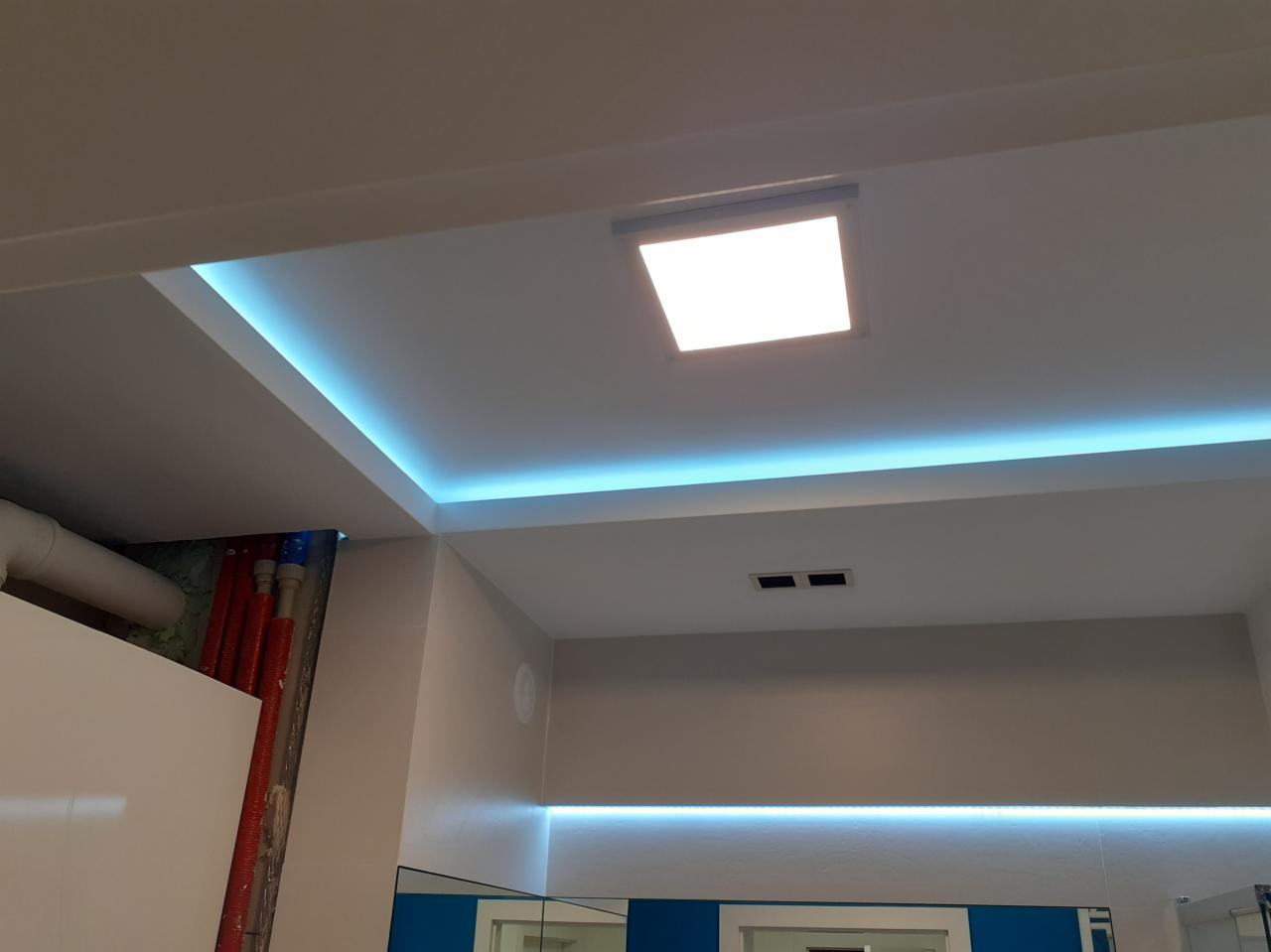 Nowoczesna łazienka z podwieszanym sufitem i oświetleniem LED w kolorze turkusowym, widoczne rury instalacyjne nad szafką, minimalistyczny styl.