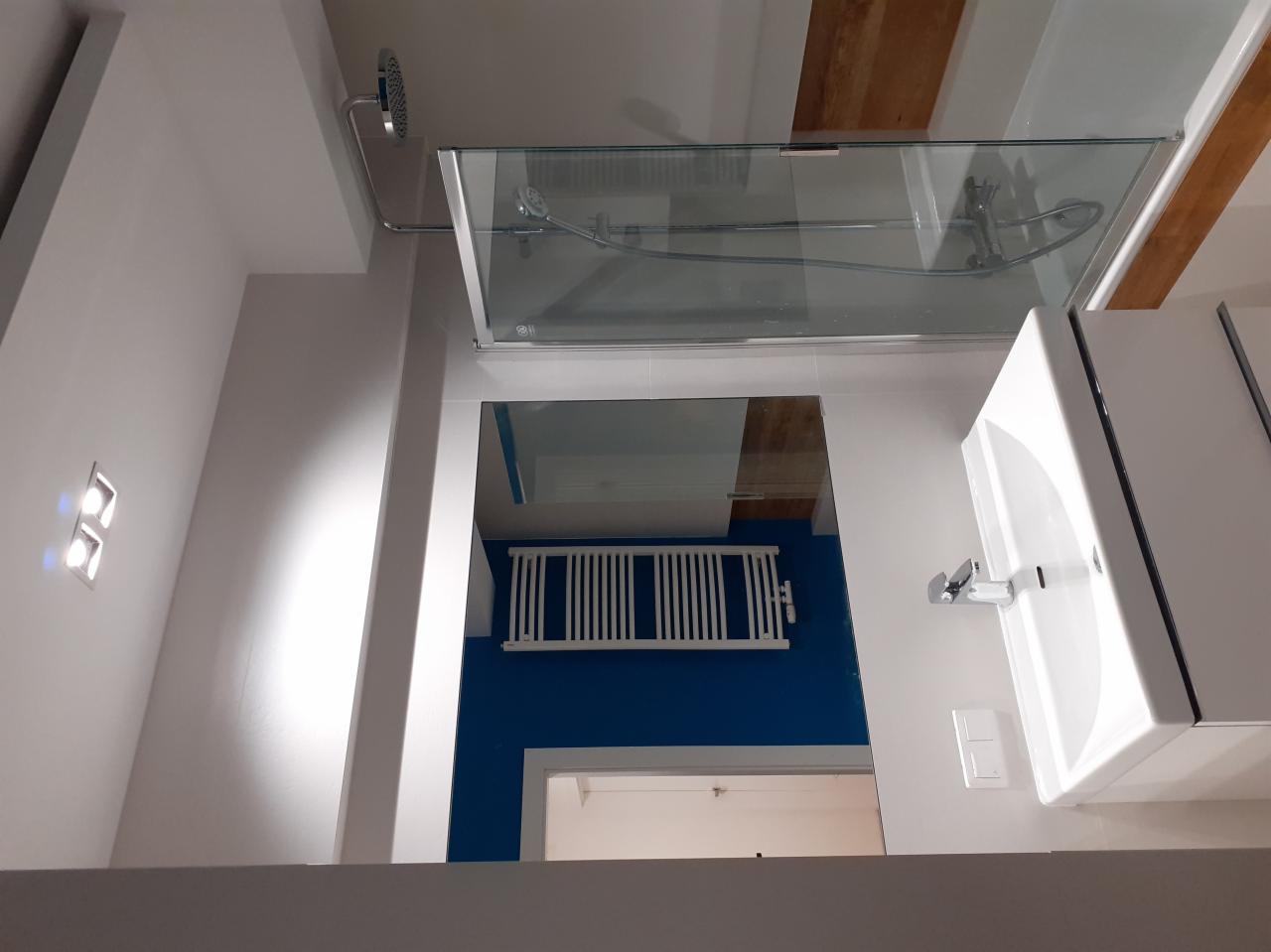 Nowoczesna łazienka z kabiną prysznicową ze szklanymi drzwiami przesuwnymi, białą umywalką z szafką i grzejnikiem drabinkowym na tle ściany pomalowanej na intensywny niebieski kolor, widoczne...