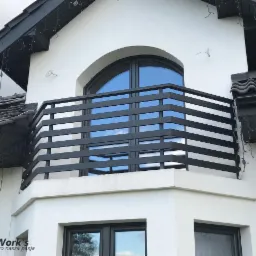 Nowoczesna, czarna balustrada balkonowa z poziomych profili, zamontowana na białym budynku z oknem łukowym, widok z dołu.