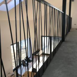 Steel-works Piotr Kowalczyk - Czarna balustrada z pionowymi prętami o nieregularnym rozstawie, widok z góry na salon z meblami, logo firmy w górnym rogu.