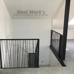 Steel-works Piotr Kowalczyk - Nowoczesna, czarna barierka schodowa z pionowymi prętami o nieregularnym rozmieszczeniu, widoczna na tle białych ścian i surowego betonowego podłoża w nowo wykończonym wnętrzu.