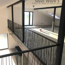 Steel-works Piotr Kowalczyk - Wnętrze domu w trakcie budowy z czarną, metalową balustradą na antresoli, widoczne elementy konstrukcyjne stalowe i butelka z płynem na podłodze.