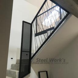 Steel-works Piotr Kowalczyk - Nowoczesna klatka schodowa z czarną balustradą o geometrycznym wzorze z cienkich prętów i panelem z perforowanej blachy, widoczny fragment betonowego stropu i logo firmy na ścianie.