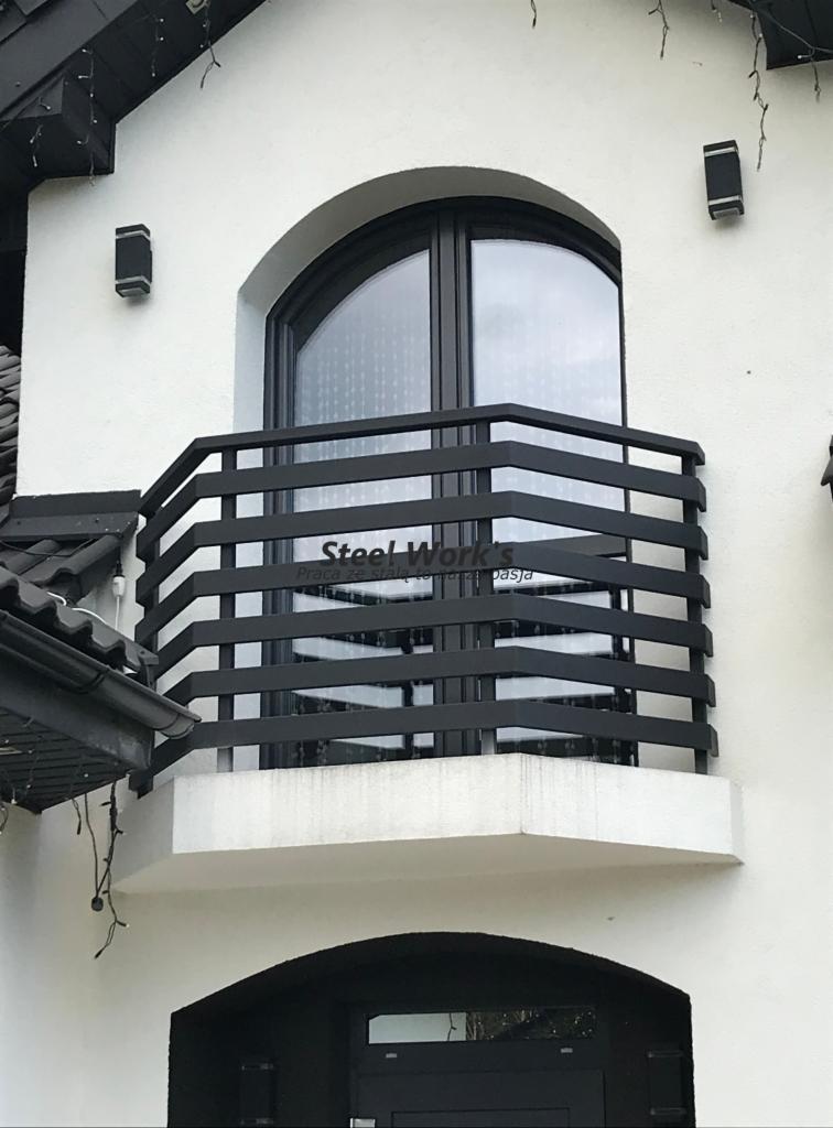 Czarna, metalowa balustrada balkonowa z poziomymi szczeblami, zamontowana na balkonie z oknem w kształcie łuku w białym budynku. Widoczne oświetlenie zewnętrzne.