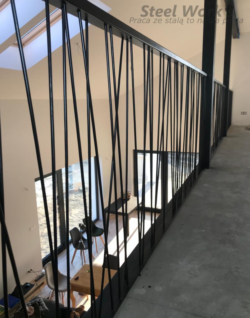 Czarna balustrada z pionowymi prętami o nieregularnym rozstawie, widok z góry na salon z meblami, logo firmy w górnym rogu.