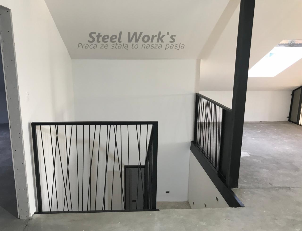 Nowoczesna, czarna barierka schodowa z pionowymi prętami o nieregularnym rozmieszczeniu, widoczna na tle białych ścian i surowego betonowego podłoża w nowo wykończonym wnętrzu.