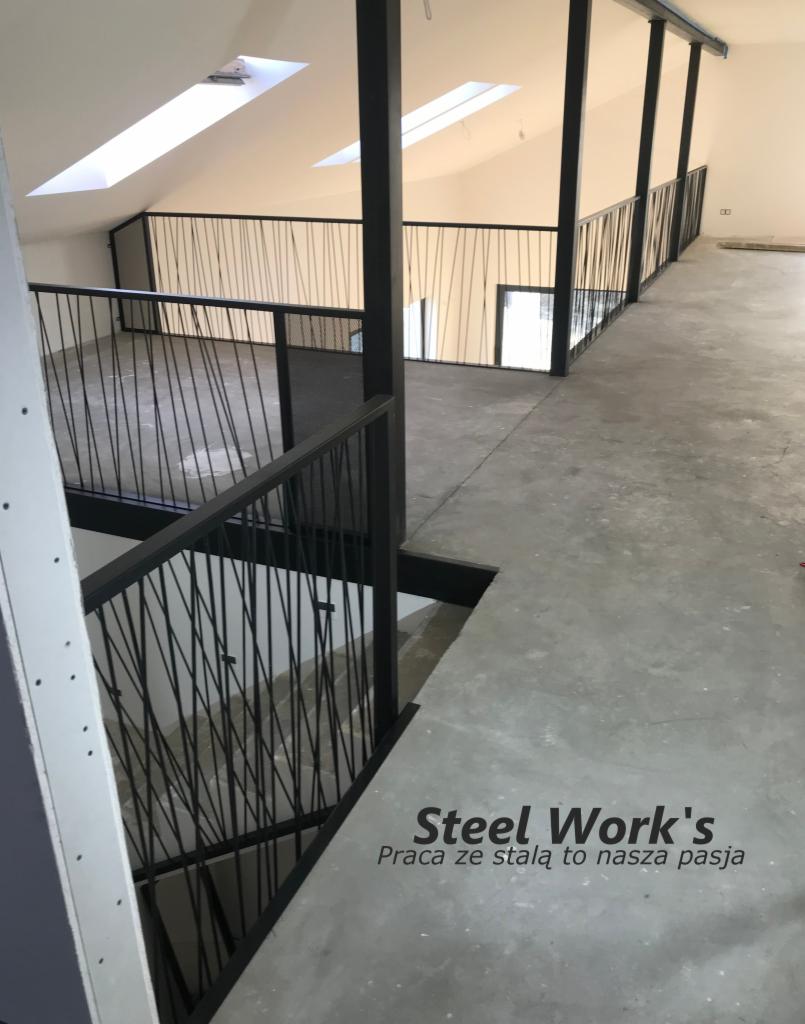 Wnętrze poddasza z czarnymi, stalowymi balustradami o minimalistycznym designie, widok z góry na schody i korytarz, logo firmy na posadzce.