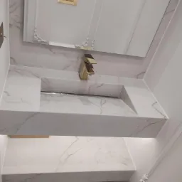 Elegancka umywalka z białego marmuru z złotą armaturą, zamontowana w nowoczesnej łazience z lustrem w ozdobnej ramie.