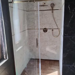 Szklana kabina prysznicowa z przesuwnymi drzwiami, złote wykończenia, marmurowe płytki na ścianach, drewnopodobna podłoga, czarne marmurowe płytki na bocznej ścianie.