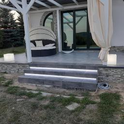 Usługi remontowo- budowlane Przemysław Bąk - Zadaszony taras z szarych płytek z podświetlanymi schodami, okrągłym siedziskiem z baldachimem i lampionami w kształcie kostek.