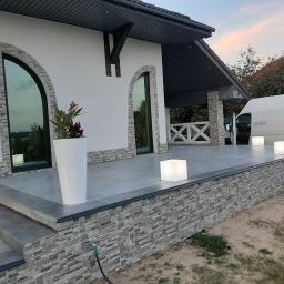 Usługi remontowo- budowlane Przemysław Bąk - Wykończony taras domu z szarymi płytkami i dekoracyjną elewacją z kamienia, z białymi donicami z kwiatami i podświetlanymi kostkami oraz zaparkowanym białym vanem w tle.