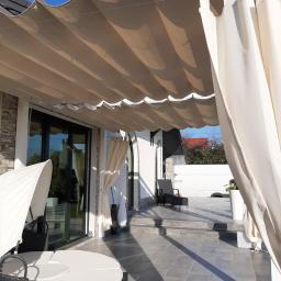 Usługi remontowo- budowlane Przemysław Bąk - Zadaszony taras z przesuwną markizą w odcieniach beżu i kremowymi zasłonami, okrągła leżanka z baldachimem, szare płytki podłogowe, widok na ogród i błękitne niebo.