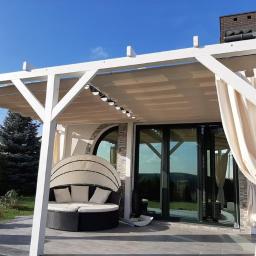Usługi remontowo- budowlane Przemysław Bąk - Biała pergola z beżową tkaniną zacieniającą i zwiewnymi zasłonami, przylegająca do budynku z dużymi przesuwnymi drzwiami i okrągłym oknem, na kamiennej posadzce, z okrągłą leżanką z poduszkami...