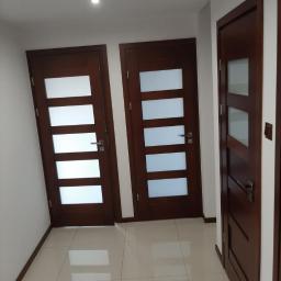 Usługi remontowo- budowlane Przemysław Bąk - Wnętrze korytarza z trzema zamontowanymi, nowoczesnymi drzwiami z ciemnego drewna, posiadającymi przeszklenia w postaci poziomych pasów, widoczne na tle jasnych ścian i podłogi z płytek.