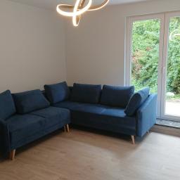 Usługi remontowo- budowlane Przemysław Bąk - Nowoczesny narożnik w odcieniach granatu na drewnianej podłodze, z widokiem na zielony ogród przez białe, dwuskrzydłowe drzwi balkonowe z marmurowym progiem, oświetlony designerską lampą sufitową.