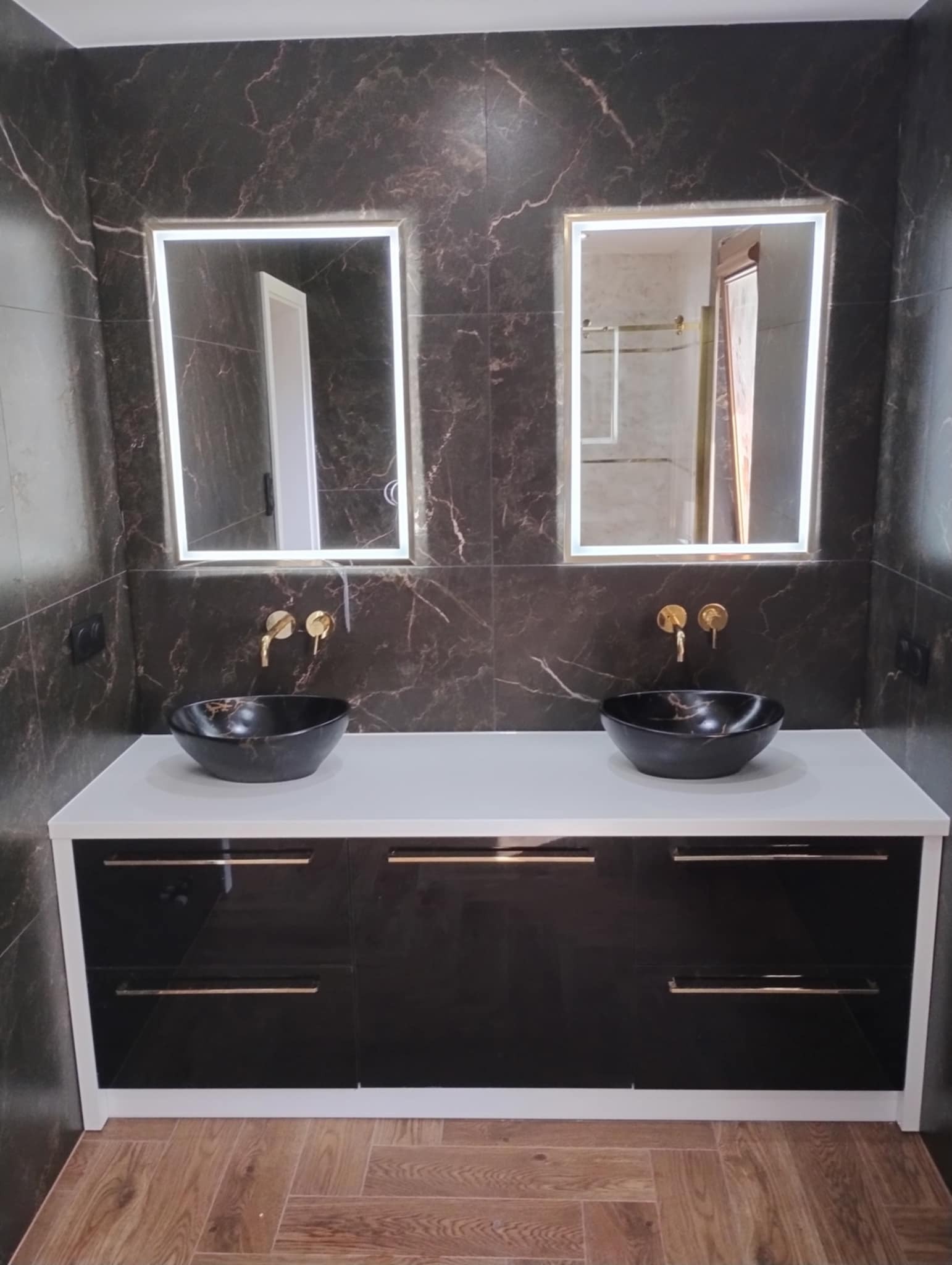 Nowoczesna łazienka z czarnymi umywalkami nablatowymi, złotą armaturą i podświetlanymi lustrami na tle płytek imitujących ciemny marmur, uzupełniona szafką z czarnymi, lakierowanymi frontami...