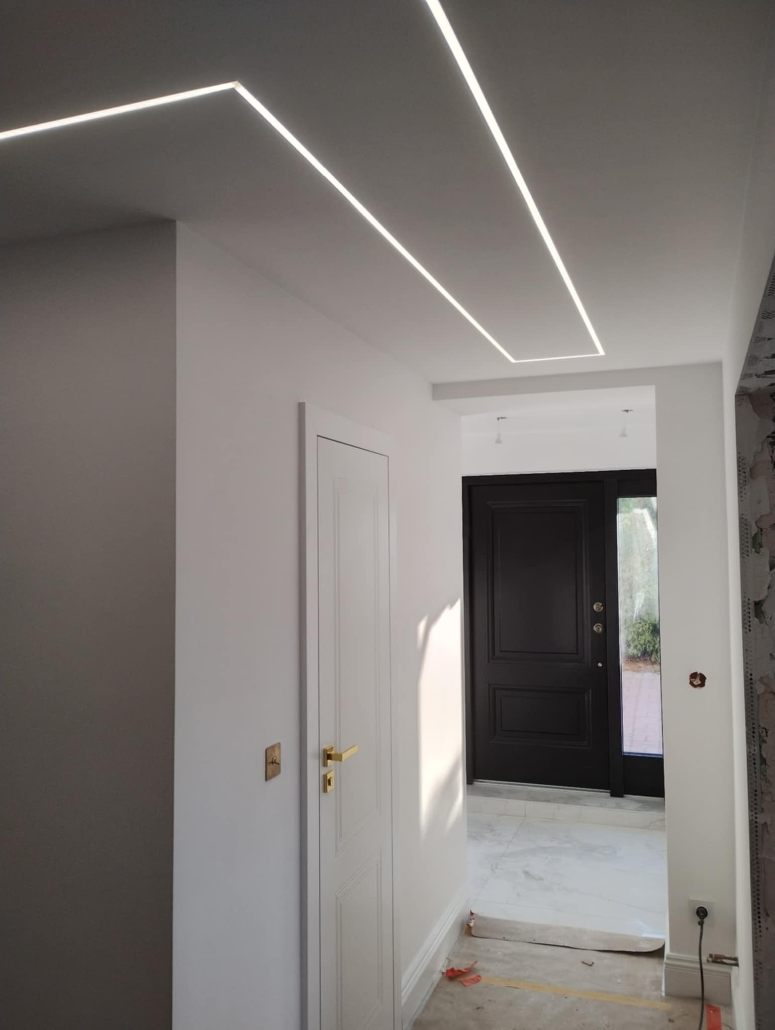 Korytarz z białymi ścianami i sufitem, oświetlony trzema liniowymi lampami LED wpuszczonymi w sufit, widoczne białe drzwi z złotą klamką oraz czarne drzwi wejściowe w tle.