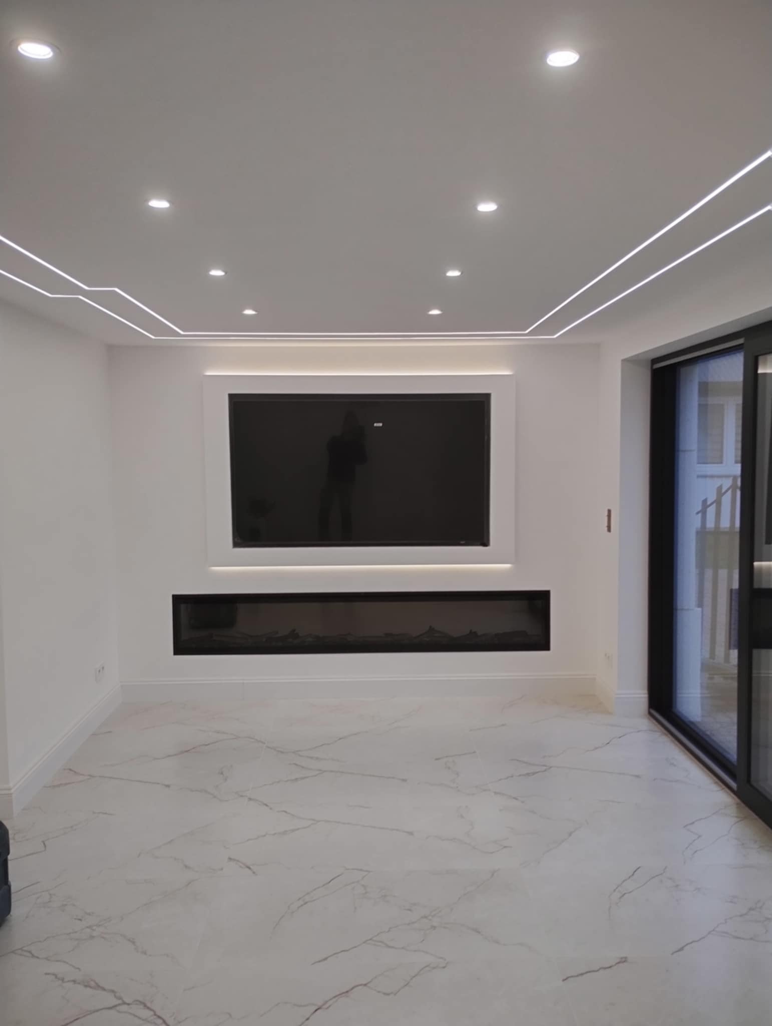 Salon z białymi ścianami, marmurową podłogą, telewizorem i elektrycznym kominkiem, podświetlony listwami LED na suficie i ścianach.