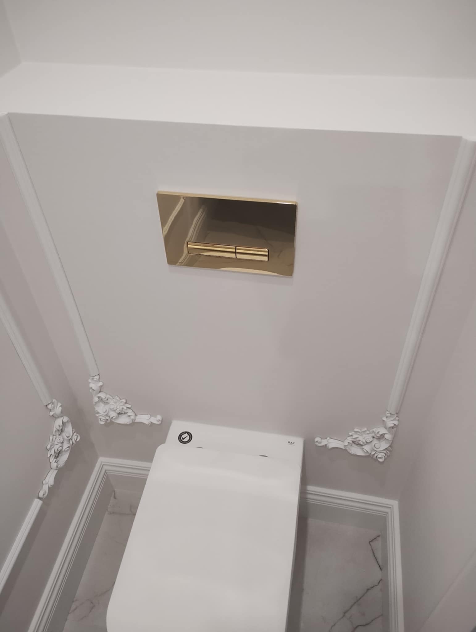 Elegancka toaleta w łazience z białymi ścianami zdobionymi sztukaterią i złotym przyciskiem spłuczki, widok z dołu.