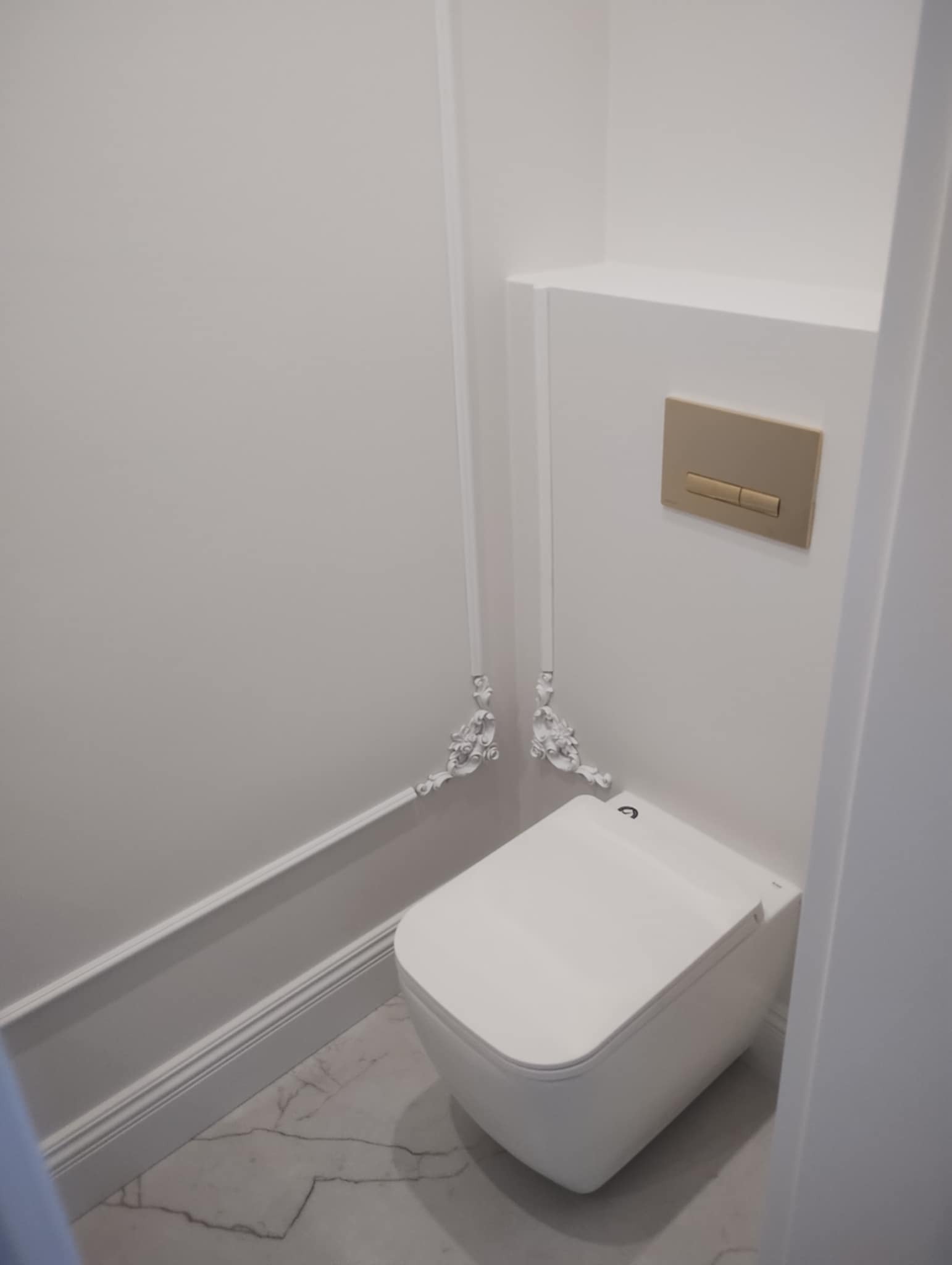 Nowoczesna toaleta podwieszana z białym wykończeniem, ozdobnymi białymi listwami przypodłogowymi i dekoracyjnymi elementami przy rurach, na podłodze marmurowe płytki, przycisk spłuczki w kolorze...