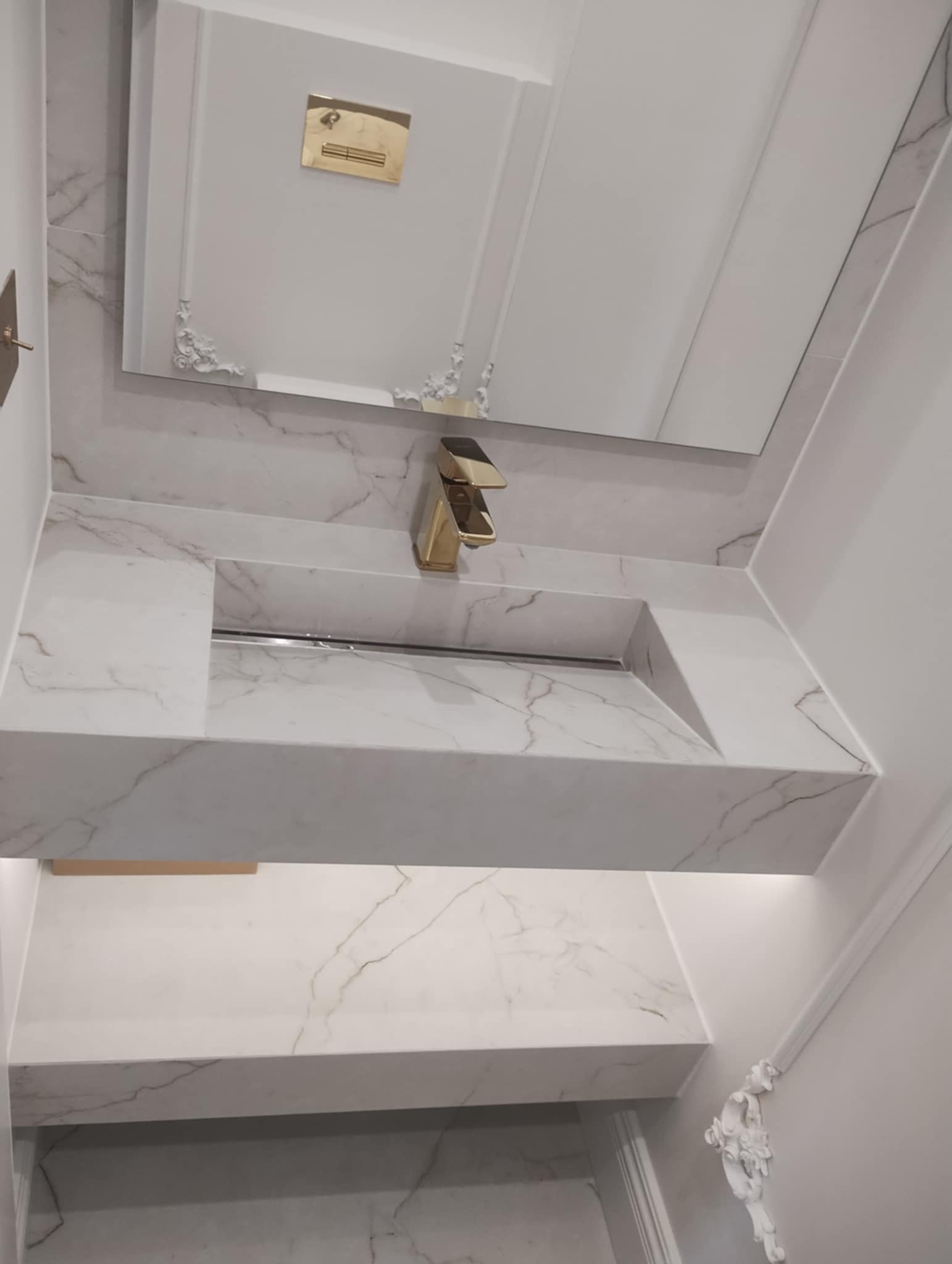 Elegancka umywalka z białego marmuru z złotą armaturą, zamontowana w nowoczesnej łazience z lustrem w ozdobnej ramie.