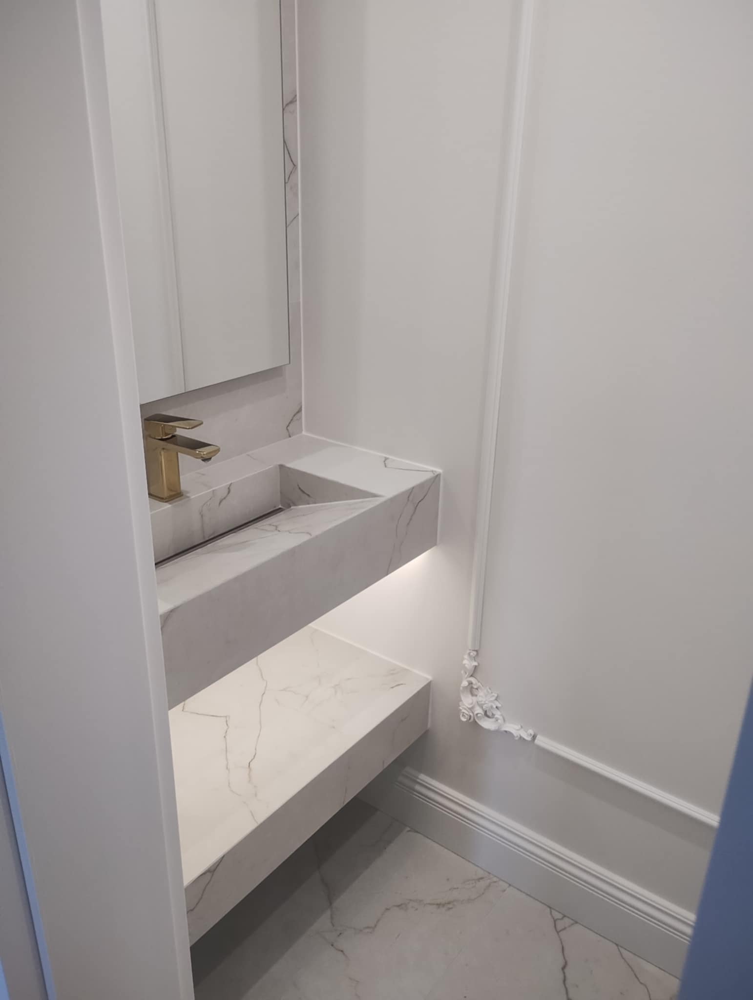 Nowoczesna umywalka z białego marmuru z podświetleniem LED i złotą armaturą, wkomponowana w minimalistyczne, białe wnętrze łazienki z dekoracyjną sztukaterią.