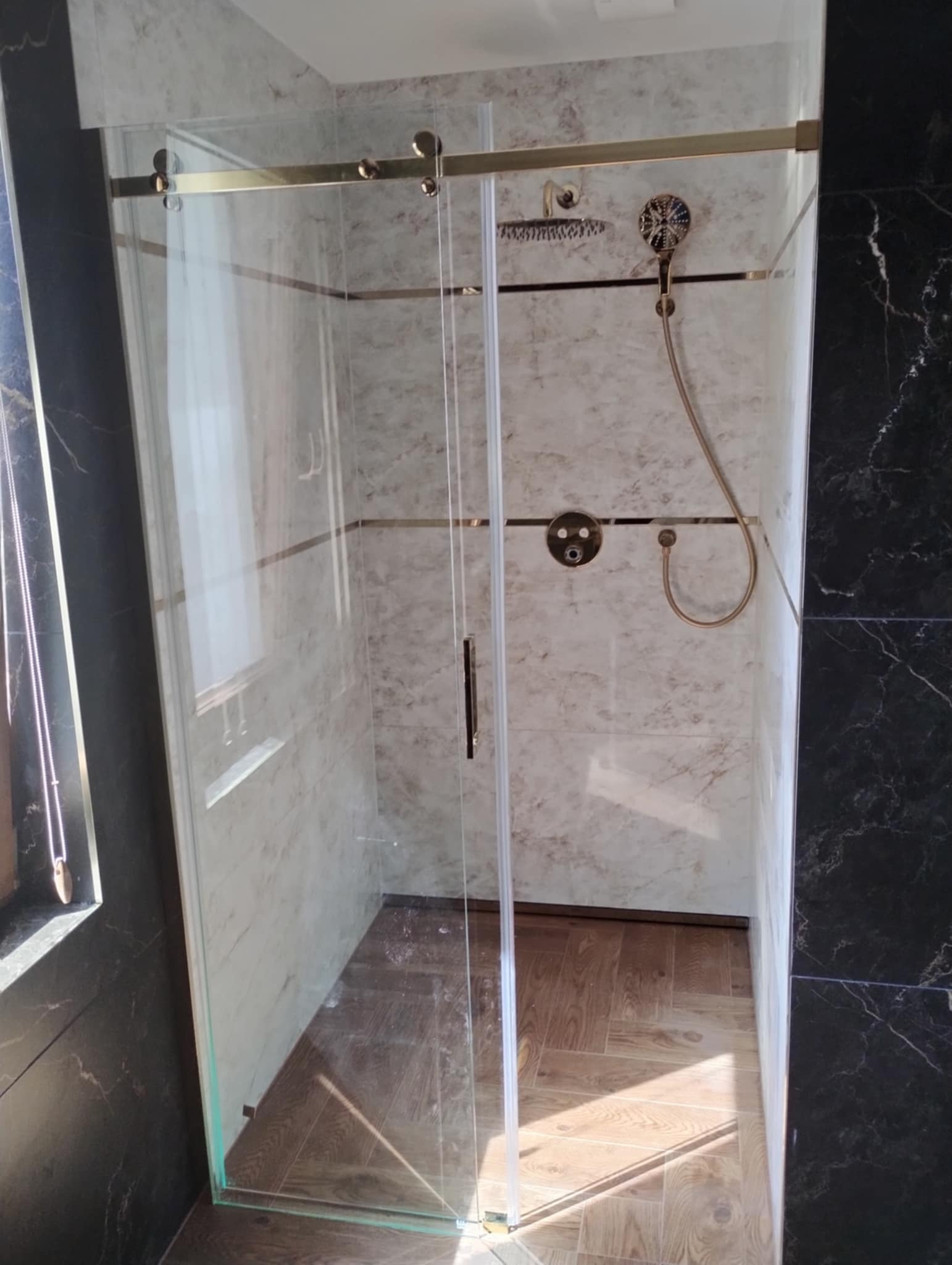 Szklana kabina prysznicowa z przesuwnymi drzwiami, złote wykończenia, marmurowe płytki na ścianach, drewnopodobna podłoga, czarne marmurowe płytki na bocznej ścianie.