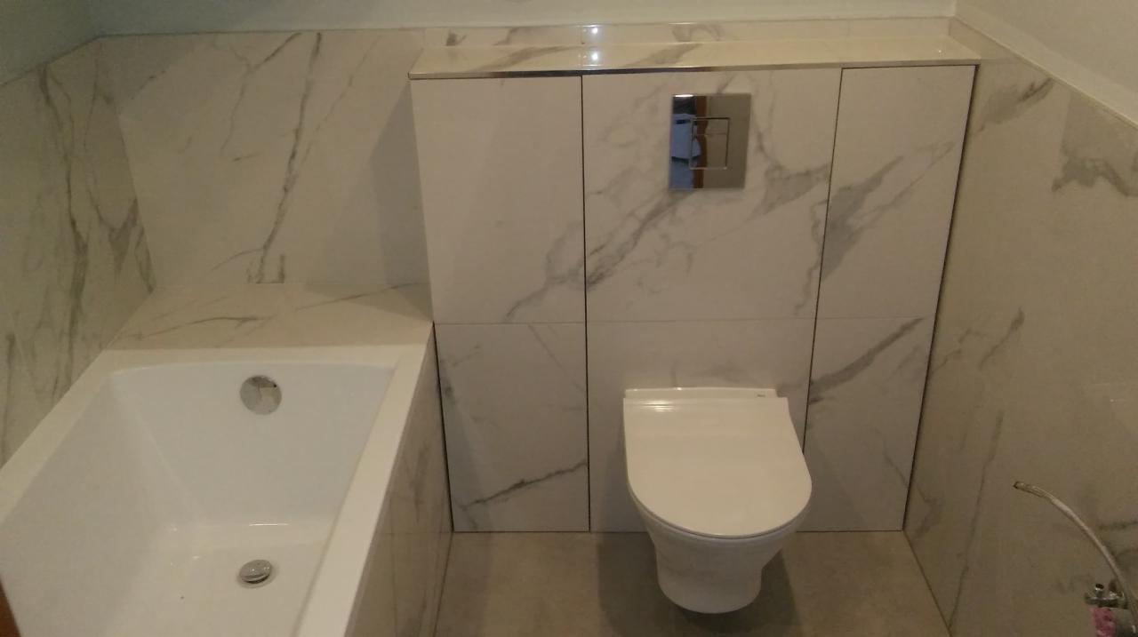 Nowoczesna łazienka z wanną i podwieszaną toaletą, wykończona białymi płytkami imitującymi marmur z szarymi żyłami, zabudowa nad toaletą z ukrytym systemem spłukiwania.