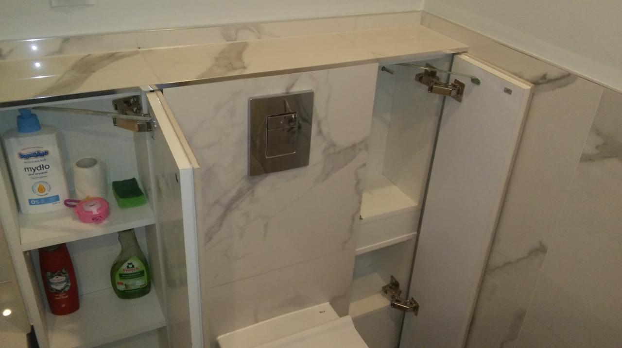 Wykończona łazienka z zabudowaną toaletą i szafkami, widoczne marmurowe płytki, chromowany przycisk spłuczki i otwarte szafki z kosmetykami.