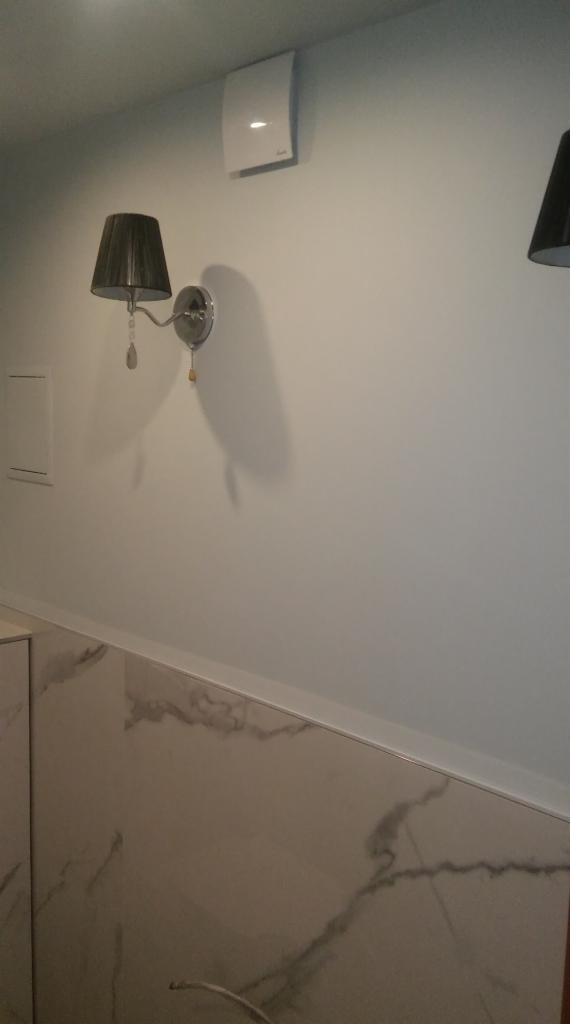 Ściana łazienki z marmurowymi płytkami, kinkietem z czarnym abażurem i kwadratowym wentylatorem na górze, widoczne delikatne oświetlenie.