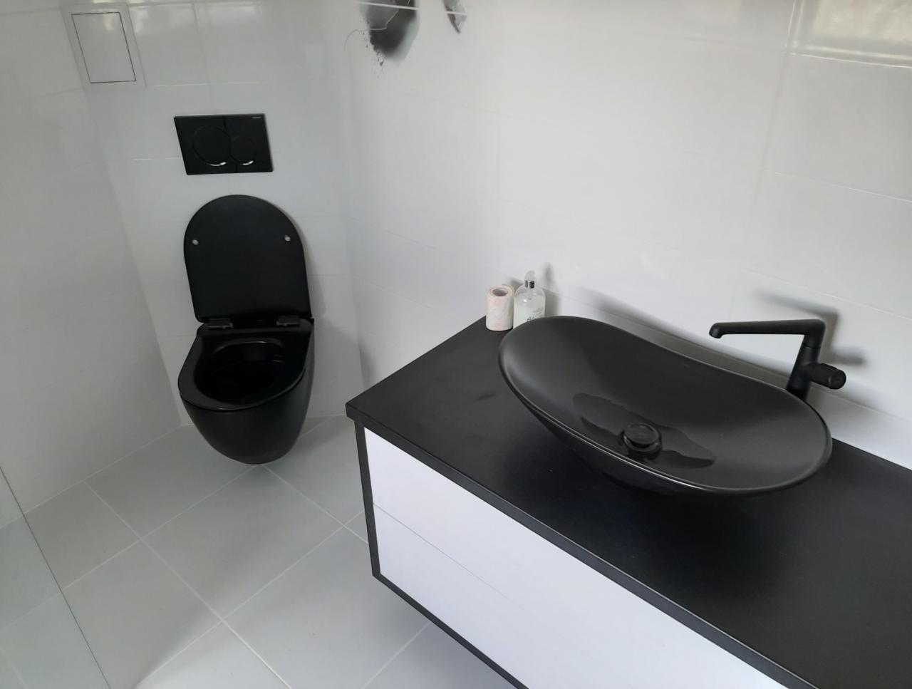Nowoczesna łazienka z wiszącą, czarną toaletą i owalną umywalką w kolorze czarnym na blacie z białymi szufladami, białe płytki na ścianach i podłodze.
