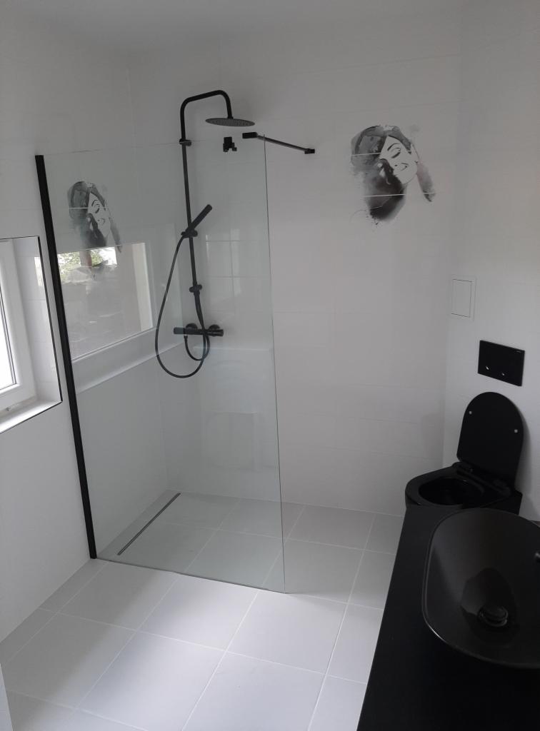 Nowoczesna łazienka z czarną armaturą: kabina prysznicowa walk-in z czarnym profilem, czarny zestaw toaletowy i umywalka nablatowa, białe płytki i dekoracja ścienna z motywem twarzy.