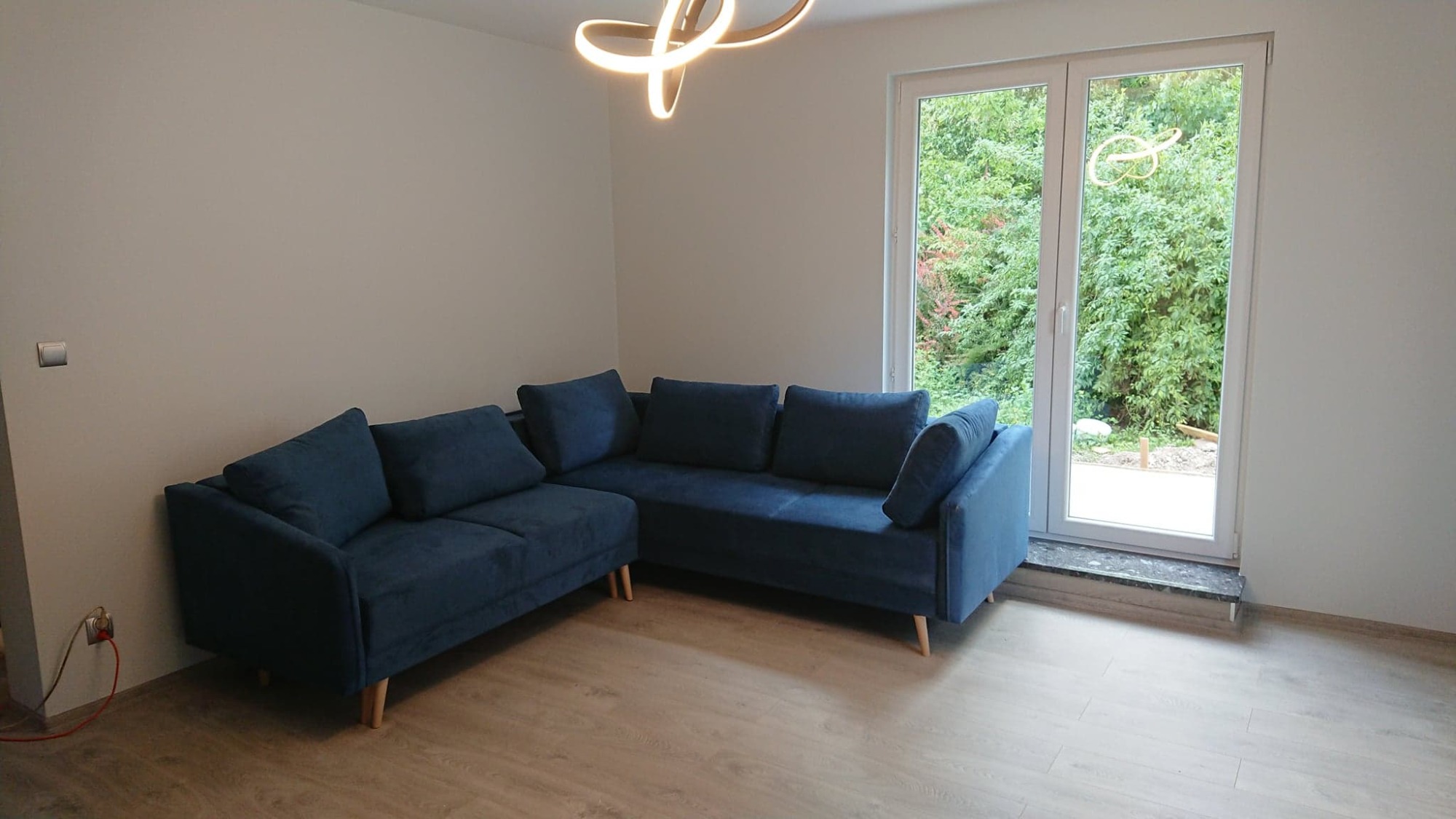 Nowoczesny narożnik w odcieniach granatu na drewnianej podłodze, z widokiem na zielony ogród przez białe, dwuskrzydłowe drzwi balkonowe z marmurowym progiem, oświetlony designerską lampą sufitową.