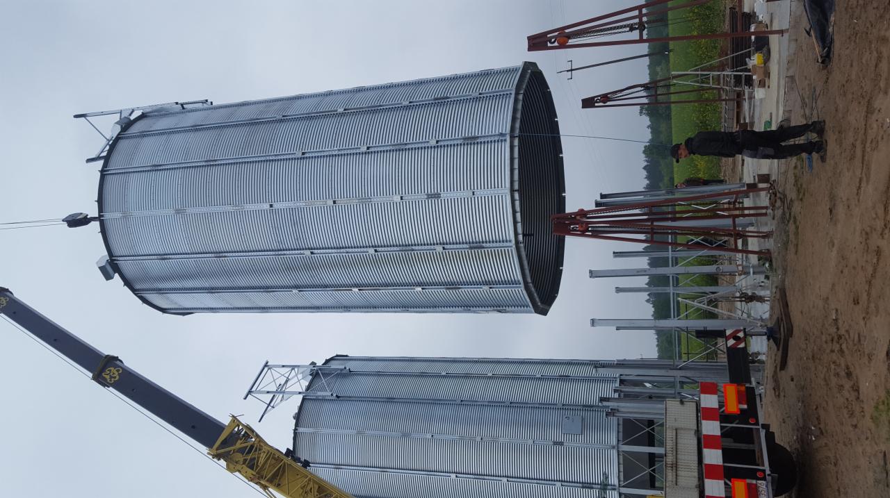 Montaż segmentu metalowego silosu zbożowego za pomocą dźwigu na placu budowy, widoczny pracownik w ciemnej odzieży.