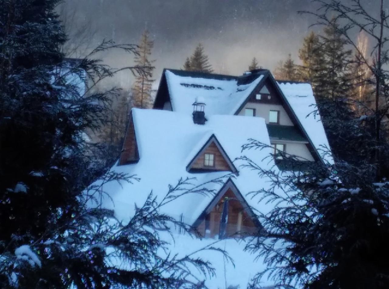 Zimowy krajobraz z ośnieżonym domem w stylu góralskim, z widocznymi spadzistymi dachami pokrytymi grubą warstwą śniegu, kominem i oknami, otoczonymi iglastymi drzewami.