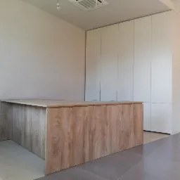 Duży, drewnopodobny blat roboczy z szafkami w tle, całość w surowym wnętrzu z szarą podłogą.