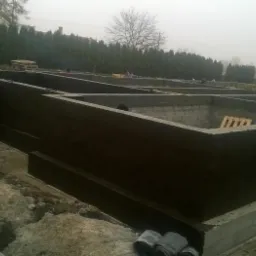 Zabezpieczone hydroizolacją fundamenty budynku w trakcie budowy, widoczne bloczki fundamentowe i izolacja pionowa, w tle zieleń i pochmurne niebo.