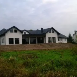 Nowoczesny dom z białymi ścianami i ciemnym dachem, duże okna, teren wokół domu jeszcze niezagospodarowany, widoczne pozostałości po budowie.