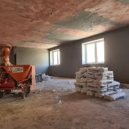Wnętrze w trakcie prac wykończeniowych: ściany pokryte świeżym tynkiem, widoczna maszyna tynkarska Malek M-4 MAX, stos worków z zaprawą oraz narzędzia budowlane. Sufit częściowo otynkowany,...