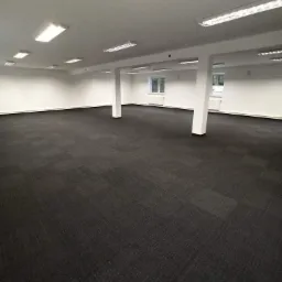 Puste, odświeżone wnętrze biurowe z ciemną wykładziną dywanową, białymi ścianami i kolumnami oraz rzędem lamp sufitowych.