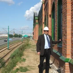 Mężczyzna w kasku i garniturze stoi na tle ceglanego budynku z zielonymi akcentami, obok torów kolejowych i autostrady.