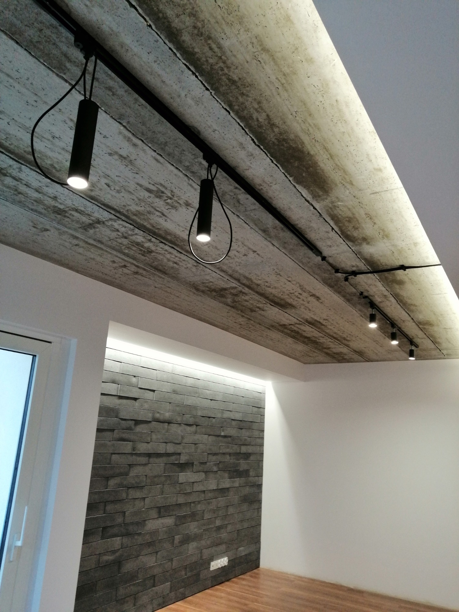Betonowy sufit z zamontowanym systemem szynowym oświetlenia punktowego, ściana wykończona szarymi płytkami imitującymi kamień, podświetlenie LED w narożniku, drewniana podłoga.
