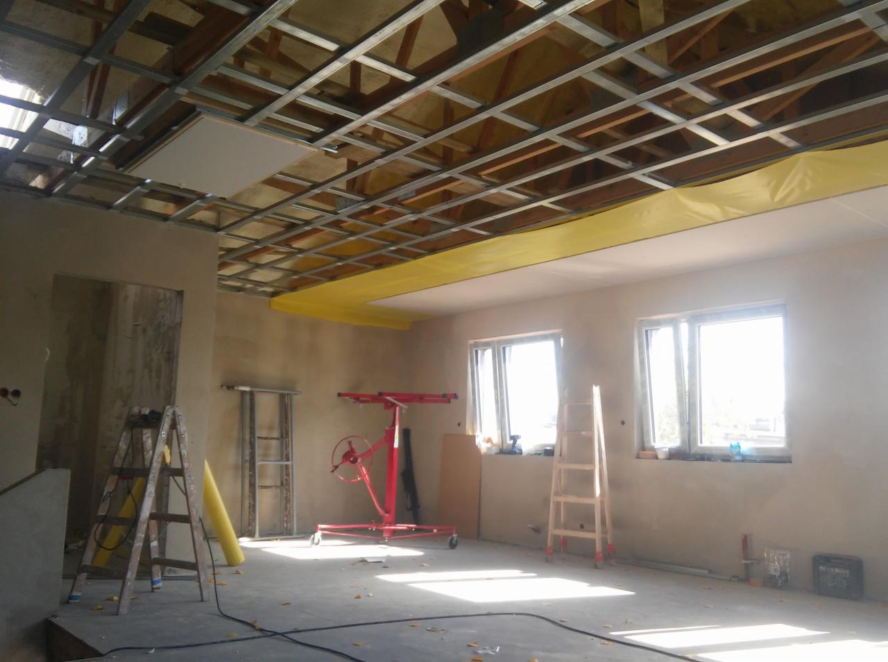 Wnętrze w trakcie prac: metalowa konstrukcja sufitu podwieszanego z zamontowaną częścią płyty kartonowo-gipsowej, zabezpieczenie folią, podnośnik do płyt, drabiny i narzędzia.