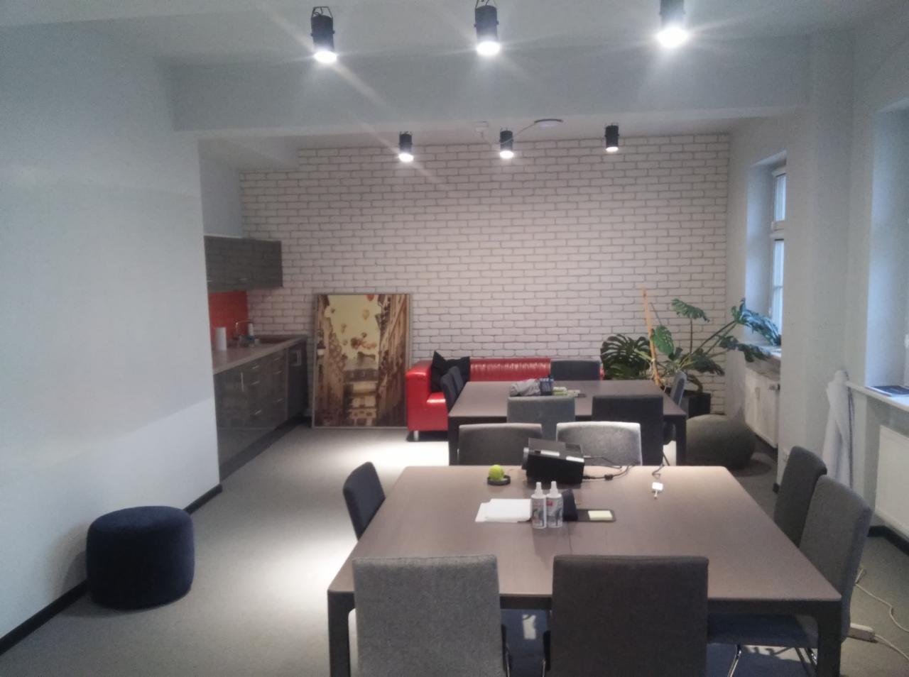 Jasne biuro z aneksem kuchennym, białymi cegłami na ścianie, drewnianymi stołami i szarymi krzesłami, oświetlone punktowymi lampami sufitowymi i światłem dziennym z okna.