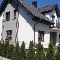 Nowoczesny dom jednorodzinny z biało-szarą elewacją, ciemną dachówką i balkonem z szklaną balustradą, widoczny przez metalowe ogrodzenie z tujami.